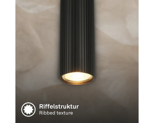Spot de plafond cylindrique noir avec une structure nervurée distinctive et une source lumineuse blanc chaud allumée.