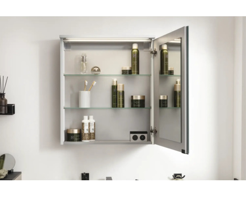 Armoire de toilette dans la salle de bains avec éclairage, porte ouverte, tablettes en verre et prise intégrée, garnie de produits de soin.