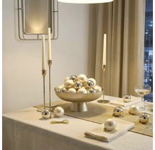 Table décorée avec une coupe sur pied remplie de boules de Noël, de hauts chandeliers et des serviettes dans une pièce lumineuse.