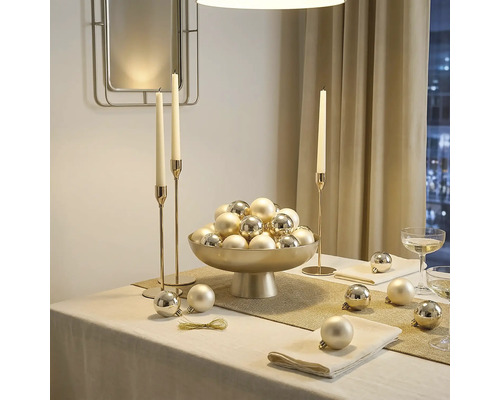 Table décorée avec une coupe sur pied remplie de boules de Noël, de hauts chandeliers et des serviettes dans une pièce lumineuse.