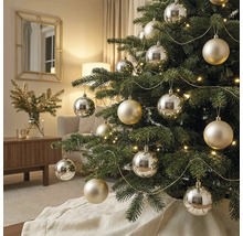Sapin de Noël décoré avec des boules argentées et couleur champagne, une guirlande lumineuse et des guirlandes dorées dans un salon lumineux.