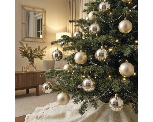 Sapin de Noël décoré avec des boules argentées et couleur champagne, une guirlande lumineuse et des guirlandes dorées dans un salon lumineux.