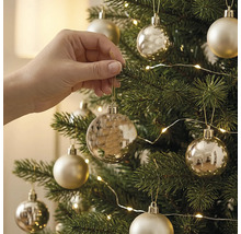 Une main suspend une boule de Noël dorée à un sapin de Noël vert décoré de lumières et d'autres décorations.