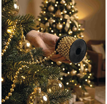 Une main tient une bobine de guirlande de perles pour décorer un sapin de Noël avec des boules et des lumières.