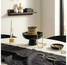 Décoration de table festive avec une guirlande de perles dorées sur une bobine dans une coupe noire, un chandelier en verre et un chemin de table.