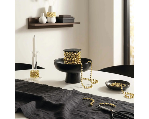 Décoration de table festive avec une guirlande de perles dorées sur une bobine dans une coupe noire, un chandelier en verre et un chemin de table.