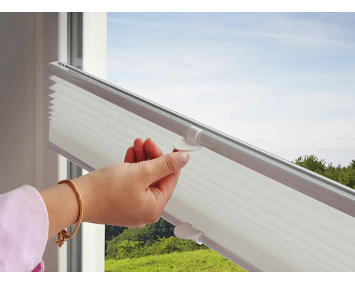 Weiße Plissee-Jalousie am Fenster, die von einer Hand am Griff nach oben geschoben wird.