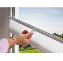 Hand bedient den Griff eines weißen Plissees an einem Fenster mit Blick ins Grüne.