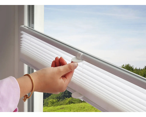 Hand bedient den Griff eines weißen Plissees an einem Fenster mit Blick ins Grüne.