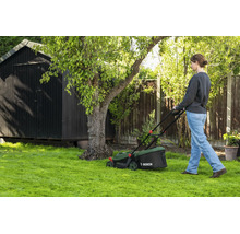 Femme tondant la pelouse avec une tondeuse Bosch dans un jardin avec un abri en bois et un arbre.