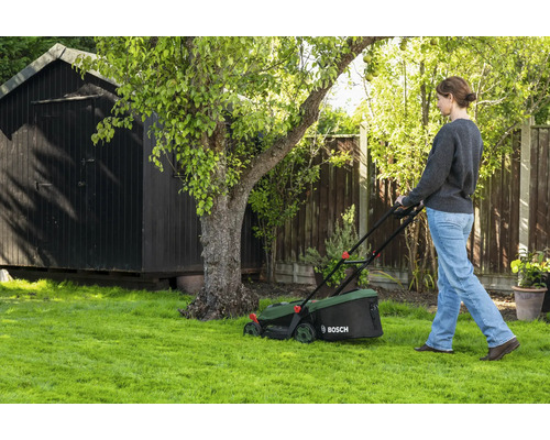 Femme tondant la pelouse avec une tondeuse Bosch dans un jardin avec un abri en bois et un arbre.