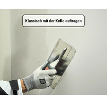 Eine Hand in einem Arbeitshandschuh trägt mit einer Glättkelle aus Edelstahl Spachtelmasse auf eine Wand auf.