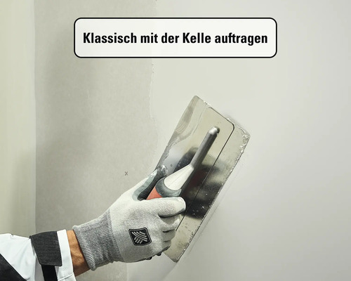 Eine Hand in einem Arbeitshandschuh trägt mit einer Glättkelle aus Edelstahl Spachtelmasse auf eine Wand auf.