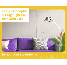 Modernes Wohnzimmer mit Wandgestaltung in Betonoptik hinter einem violetten Sofa mit Kissen und einer silbernen Lampe.