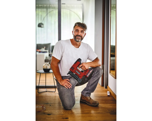 Artisan agenouillé sur un parquet tenant une agrafeuse sans fil Einhell dans un intérieur moderne.