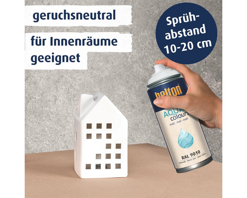Hand sprüht Belton Aqua Sprühlack auf ein weißes Deko Haus. Informationen zu Geruchsneutralität, Eignung für Innenräume und Sprühabstand.