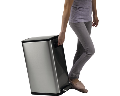 Une personne actionne la pédale d''une poubelle en acier inoxydable tout en la soulevant par la poignée.