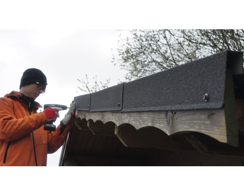 Un homme fixe du carton bitumé sur un cabanon en bois avec une visseuse sans fil.