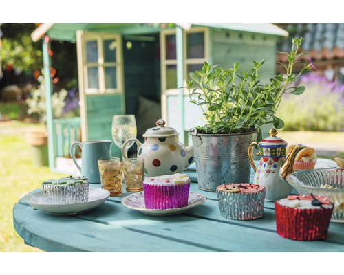 Table dressée avec cabane de jardin en arrière-plan pour une garden-party avec théières, plantes, verres et muffins.