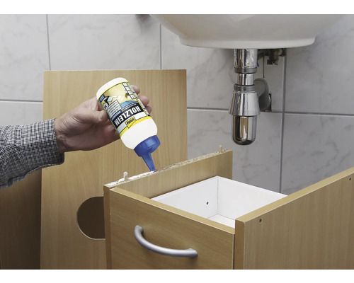 Un artisan applique de la colle à bois sur un meuble en bois dans la salle de bains. Un lavabo est visible à l''arrière-plan.
