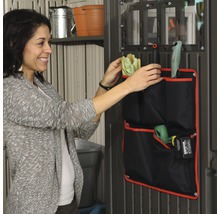 Une femme range des outils de jardin dans un sac à outils noir à plusieurs compartiments fixé au mur.