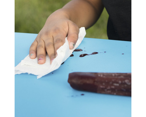 Eine Hand wischt mit einem Papiertuch eine blaue Oberfläche mit Flecken ab. Ein Eis am Stiel liegt auf dem Tisch.