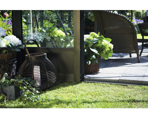 Szene mit einer Laterne, einem Korb mit Pflanzen und Gartenmöbeln auf einer Rasenfläche neben einem Gewächshaus.