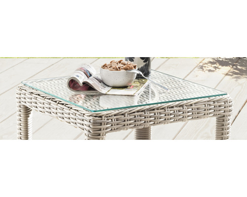 Table de jardin avec plateau en verre et structure en rotin, sur laquelle sont posés un magazine, un bol de snacks et une tasse