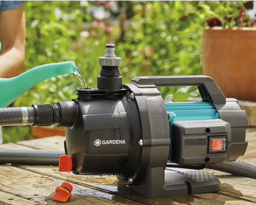 Gartenszene mit einer Gartenpumpe der Marke Gardena, die mit Wasser aus einer Gießkanne befüllt wird.