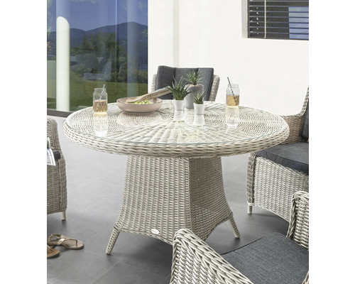 Table de jardin ronde avec plateau en verre et base en rotin, entourée de fauteuils en rotin avec coussins gris.