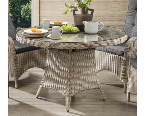 Table de jardin ronde avec plateau en verre et aspect rotin à côté de deux chaises avec coussins