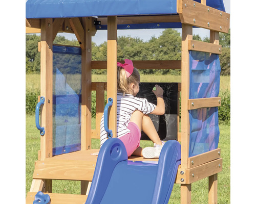 Une fille dessine sur un tableau noir dans une aire de jeux en bois avec toboggan et auvent bleu.