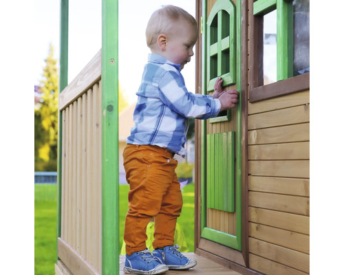Jeune enfant debout devant une maison de jeux en bois avec porte et fenêtres.