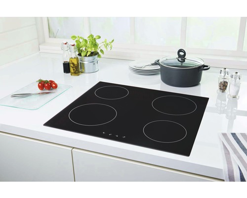 Schwarzes Glaskeramikkochfeld mit vier Kochzonen auf einer weißen Küchentheke mit Kochtopf, Tellerstapel, Gewürzmühle, Schneidebrett, Tomaten und Kräutern.