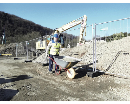 Bauarbeiter schiebt Schubkarre mit Bauschutt vor Bauzaun und Bagger.