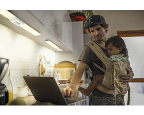 Mann mit Babytrage arbeitet am Laptop in der Küche mit Unterbauleuchten