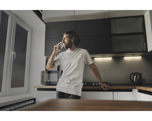 Mann trinkt ein Glas Wasser in einer Küche mit Mikrowelle, Kochfeld und Wasserkocher.