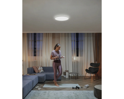 Eine Frau steht auf einer Yogamatte in einem Wohnzimmer mit einer runden Deckenleuchte.