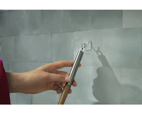 Eine Hand hängt einen Gegenstand mit einem Haken an einen transparenten Klebehaken an einer gefliesten Wand.