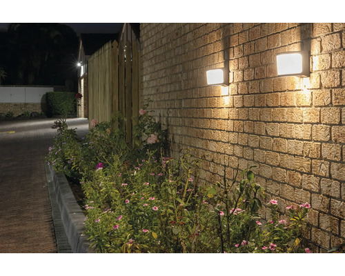 Zwei quadratische Aussenleuchten an einer Backsteinmauer neben einer Bepflanzung.