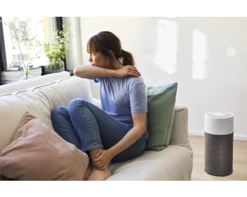 Frau hustet auf einer Couch neben einem Luftreiniger