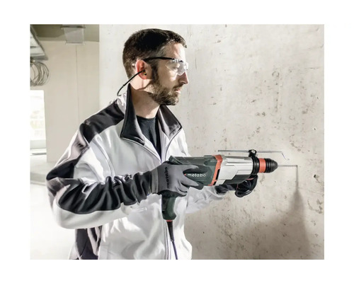 Un homme perce un mur en béton avec un perforateur. Il porte des lunettes de protection, une protection auditive et des gants.