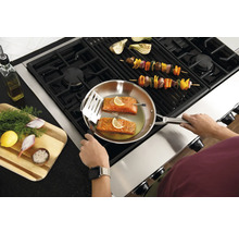 Une personne cuisine du saumon dans une poêle sur une cuisinière, tandis que des brochettes de légumes sont sur le gril.