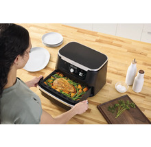 Une femme pousse une friteuse à air chaud avec du poulet et des légumes sur une table en bois avec des assiettes, des bouteilles d''huile et des herbes.
