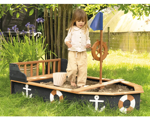 Kind spielt in einem Sandkasten in Form eines Schiffs mit Steuerrad und blauem Segel