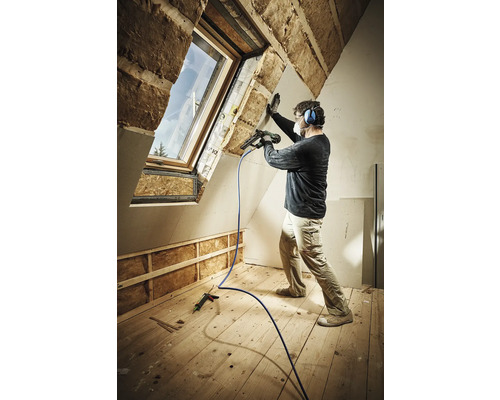 Ein Mann befestigt eine Gipskartonplatte mit einem Druckluftnagler in einem Dachgeschossraum mit Dachfenster und Holzboden. Er trägt dabei eine Atemschutzmaske und Gehörschutz.