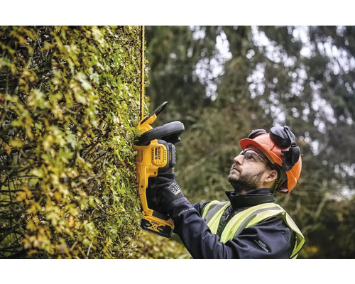 Un homme portant un casque et des lunettes de protection taille une haie avec un taille-haie sans fil DeWalt.