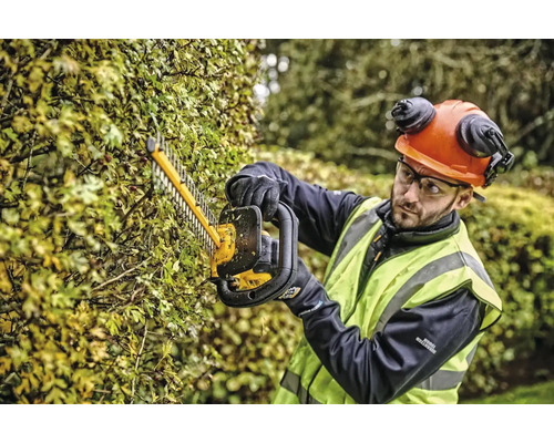 Une personne taille une haie avec un taille-haie tout en portant des vêtements de protection.