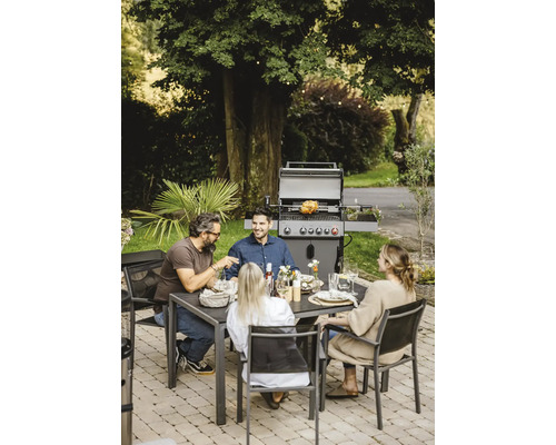 Scène extérieure avec un barbecue à gaz, une table à manger et quatre personnes en train de manger.