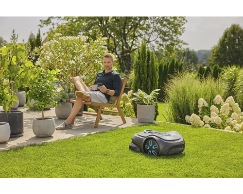 Ein Mann entspannt im Garten neben einem Rasenmähroboter, umgeben von üppiger Vegetation und Topfpflanzen.